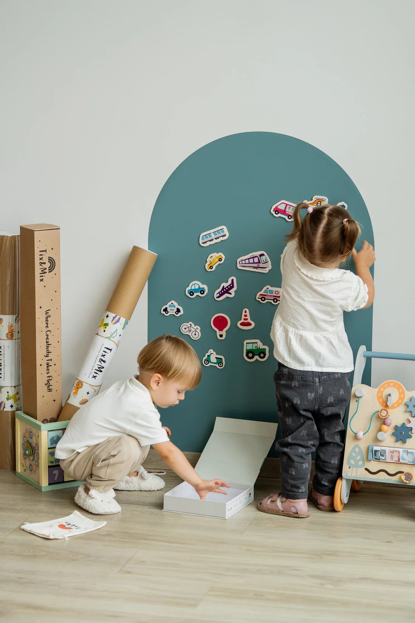 Two toddlers playing with Tix&Mix wooden magnetic vehicles playset on a cyan wave magnetic wall decal.