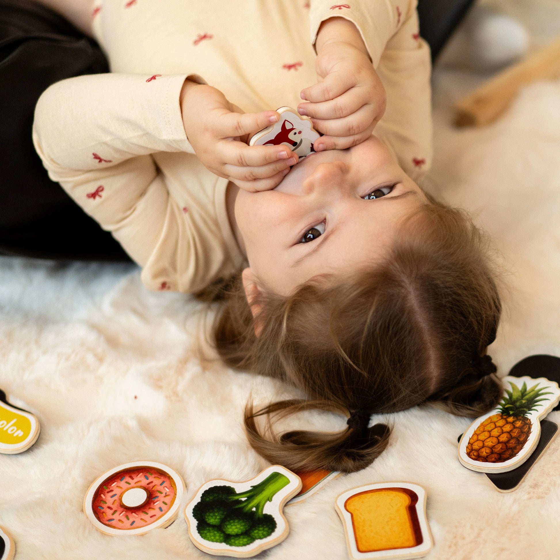 A female toddler laying on a clean mat looking at the camera while holding Tix&Mix vegetable toys set.