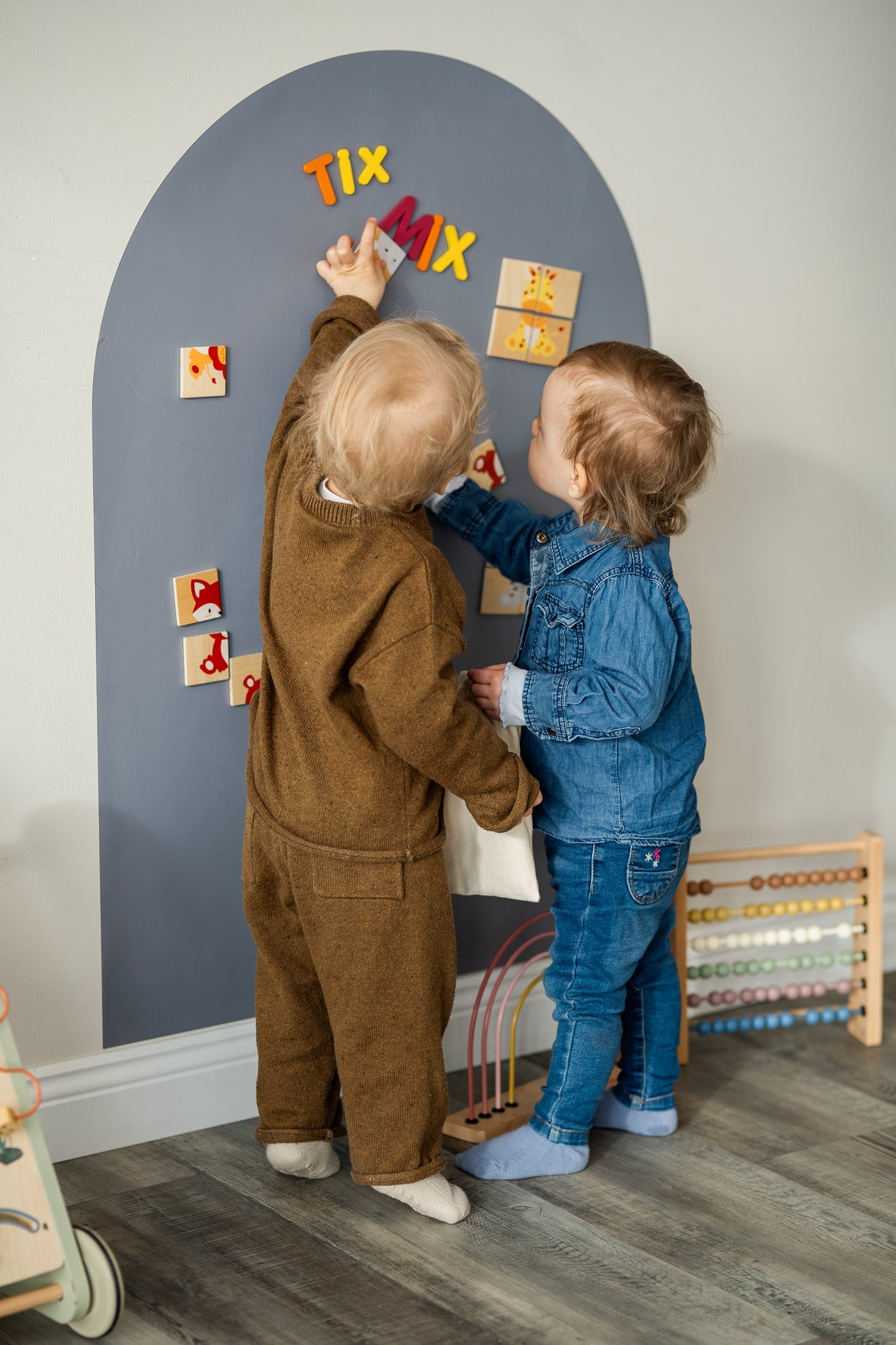 Two toddlers playing with Tix&Mix magnetic letters on a grey magnetic wall decal.