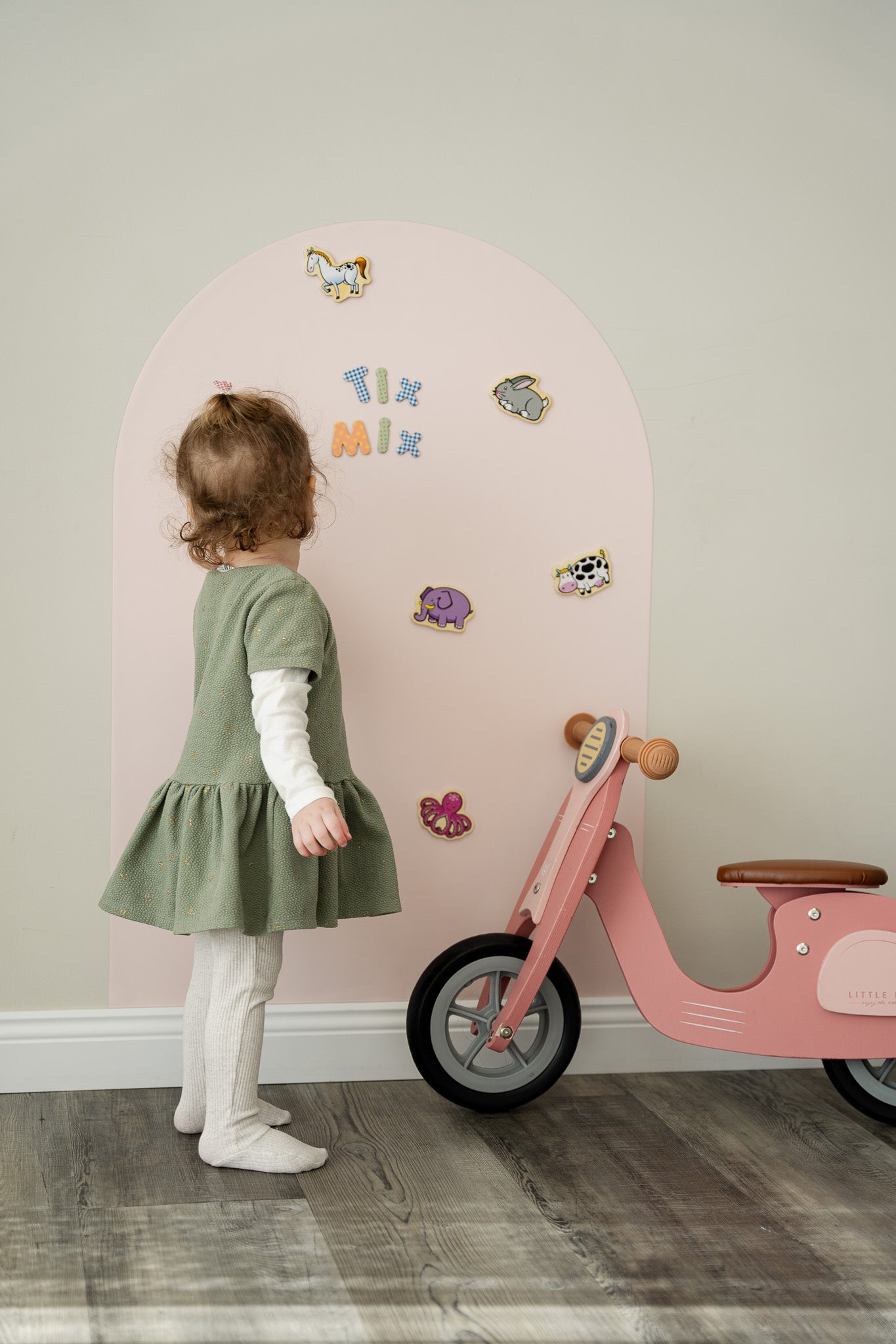 A toddler standing in front of Tix&Mix pink magnetic wall decal with magnetic toys attached to the wall decal.